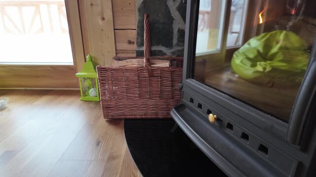 Fire glowing behind stove glass as the camera moves closer, capturing rustic reflections and a basket of prepared kindling.