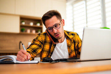 Working from home, a young man pauses to write down ideas and reminders, combining online research with traditional note-taking. © djoronimo