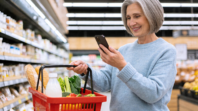 Senior woman checks phone while shopping for groceries in a store on a weekday afternoon with a basket of fresh items
