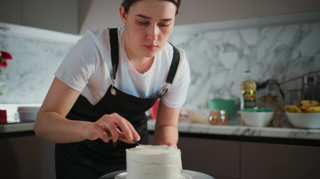 Focused young woman in an apron carefully decorates a white cake with a knife. She leans over the cake, concentrating on smoothing the frosting. The modern kitchen has marble backsplash