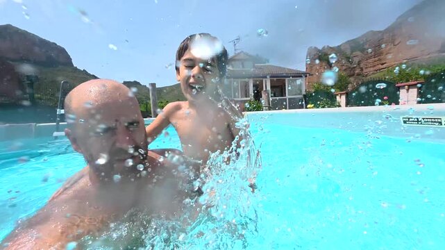 Playful son sitting on his father's shoulders and splashing water towards the camera while having fun together in a swimming pool on a sunny day, creating joyful family memories