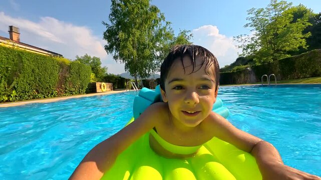 Joyful little boy having fun floating on a colorful inflatable mattress in a backyard swimming pool, smiling and laughing at the camera on a beautiful sunny day during his summer vacation