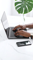 Fototapeta premium Closeup of a Black professional typing on a laptop at a bright, minimalist marble desk with a smartphone and a monstera plant. Perfect for remote work and tech concepts.