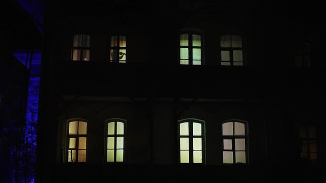 Silhouette of a man playing the violin in the evening window of a house. A persons silhouette stands in a warmly lit window of a dark house at dusk