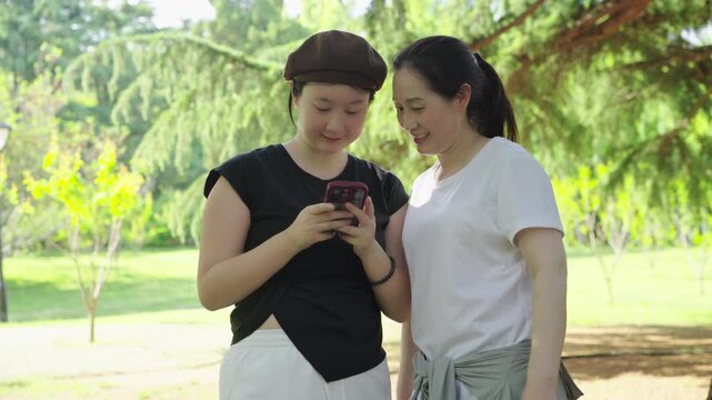 Two smiling young women stand in a sunlit park, looking at a smartphone together, with one pointing at the device screen.