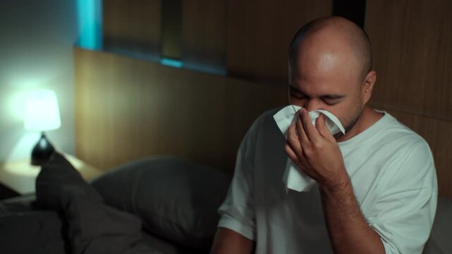 Sick man coughing and sneezing into tissue while resting on bed at home. Fever, dust allergy, respiratory illness, seasonal flu and healthcare concept.
