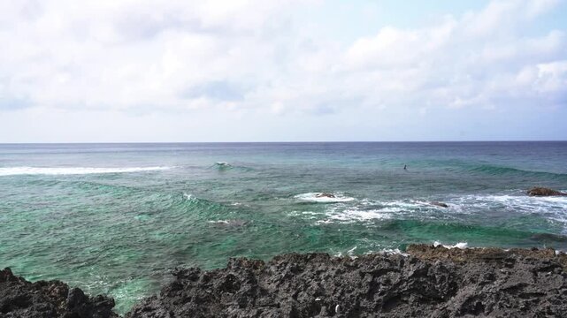 A single surfer floating on the beautiful emerald green ocean waiting for waves near a rocky coast, Yonaguni Island, Japan