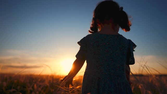 A girl stands in a field at sunset, with the sun shining brightly behind her. The girl enjoys the beautiful view of the golden field and the colorful sky.