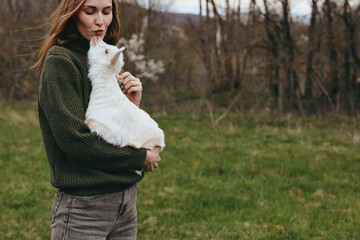 A woman in a knitted sweater cradles a white goat kid outdoors in a grassy field, sunlight filtering through trees. A gentle moment of bonding, rural life, and animal companionship in natural light. © SHOTPRIME STUDIO