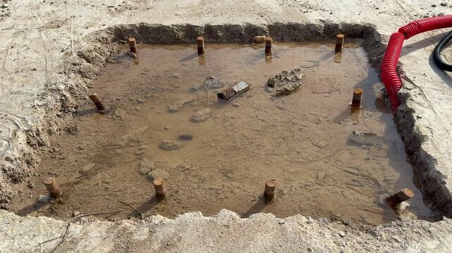 Square excavation next to coiled cable, red conduit end visible on asphalt edge, compact dirt base for bolt anchor, electrical