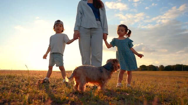 A woman stands holding hands with a boy and a girl. A small dog is nearby, all in a field during sunset, creating a warm atmosphere.