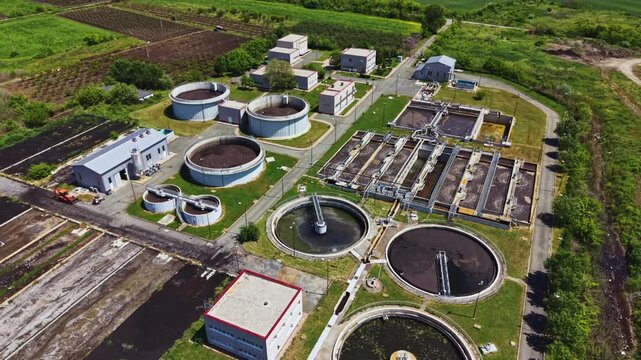 Aerial wastewater treatment plant showing circular clarifiers and sludge tanks amid green fields, service buildings and access roads industrial maintenance vibe