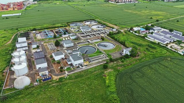 Aerial treatment plant in green fields showing full facility footprint, circular clarifiers, dome digesters, access roads, solar panels, adjacent crops,