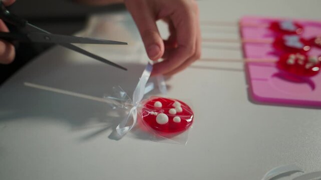Hand gently wraps a red, heart-shaped lollipop in cellophane. The lollipop features white candy dots, tied with a white ribbon. Another hand uses scissors to trim the wrapper