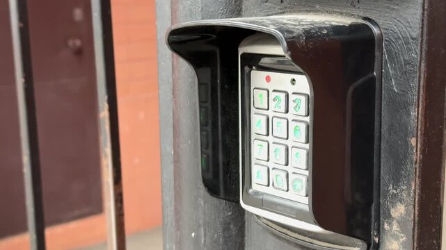 Analog intercom keypad on iron gate, weathered black frame, green backlit numeric buttons, round scanning key below keypad, brick
