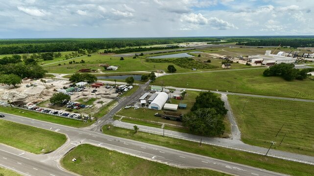 Aerial view of industrial facilities, warehouses, and parking areas surrounded by lush green fields and dense forests under a cloudy sky in Green Cove Springs, Florida, United States.