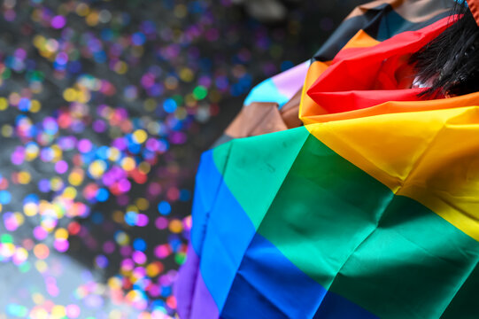 A person draped in a rainbow flag stands amidst colorful confetti on Silom Road during the Bangkok Pride Parade, a festive celebration of LGBTQ+ equality and community.