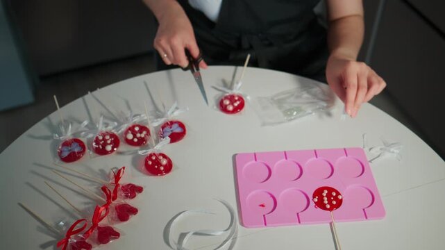 Person carefully wraps red lollipop candies with clear plastic. Pink silicone molds sit nearby. Lollipops rest on sticks, some wrapped in red ribbon. Hands cut wrapping with scissors