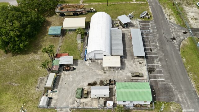 Aerial view of a military museum site with a large white arched hangar, vintage military vehicles, and outdoor exhibits under a clear sky in Green Cove Springs, Florida, United States.