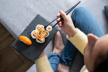 Woman eating sushi at home while holding slate serving tray sitting on the sofa © chandlervid85