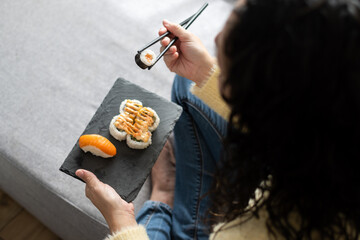 Woman eating sushi at home while holding slate serving tray sitting on the sofa © chandlervid85