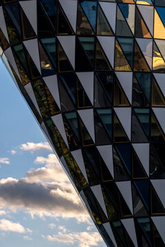Angular facade architecture leans above the street as geometric glass panels reflect sky, clouds and urban light across a patterned building.