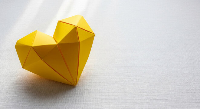 Geometric origami heart shape in bright yellow, casting shadow on textured white surface, symbolizing love, joy, and creativity.