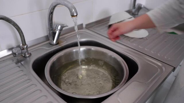 Water flows from a faucet into a stainless steel bowl in a kitchen sink while a hand reaches toward a stack of white dishes on the side
