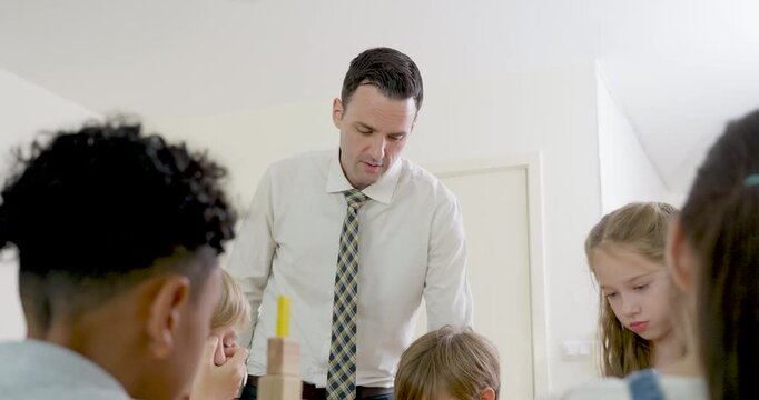 Slow motion adult male teacher supervises multicultural children during wooden block activity, guiding teamwork, participation, attention, and back to school study routine in primary classroom.