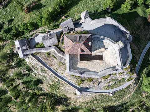 Aerial view of Schloss Tarasp featuring stone fortifications, tiled rooftops, and defensive walls surrounded by lush green forest in Scuol, Graubunden, Switzerland.