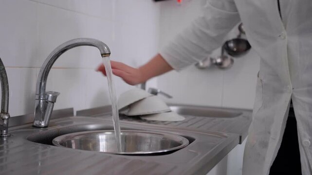 Caucasian female in white lab coat turns on faucet to fill stainless steel sink with water while washing dishes in a clean kitchen environment with utensils visible