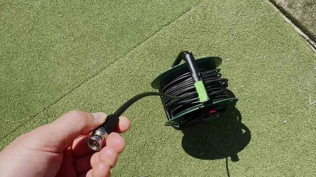 Hand holding an SDI BNC video cable connector beside a coiled cable reel on artificial turf, showing broadcast production wiring and outdoor setup detail.