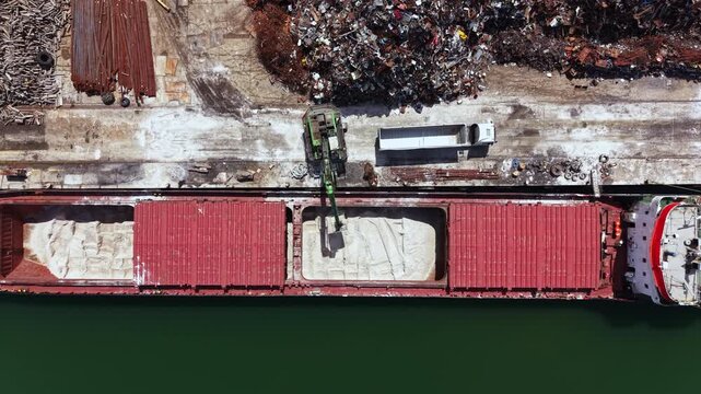 A large cargo ship is at the dock receiving sand from a loader. Scrap metal is stacked nearby, showing signs of industrial activity in a busy harbor environment.