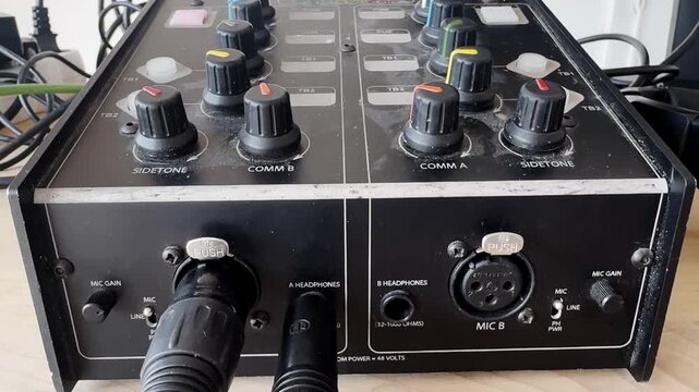 Close-up of a broadcast intercom audio control unit with comm knobs, sidetone dials, microphone inputs, headphone jacks, cables, and production equipment detail.