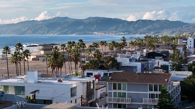Forward-rotating wide-angle aerial drone shot over Venice Beach's Ocean Front Walk, sweeping from beachside homes to palm-lined promenade with mountains beyond on a bright Los Angeles day.
