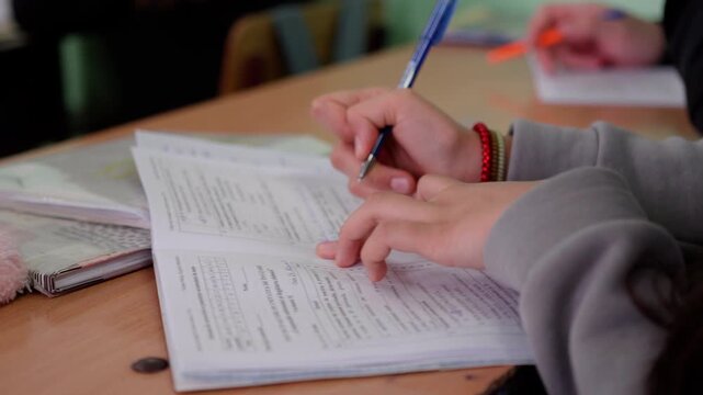 Female student with long hair writes notes in a notebook using a blue pen while seated at a desk in a classroom with other students visible in the background