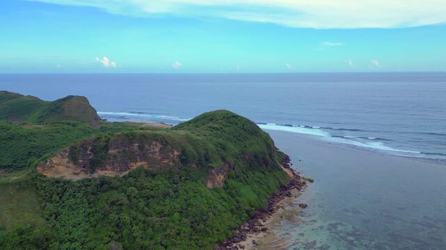 Lombok island coastline view showing lush green hills, a winding path, distant ocean waves, and clear shallow water. Marvelous aerial view speed ramp time lapse fly reverse panorama drone