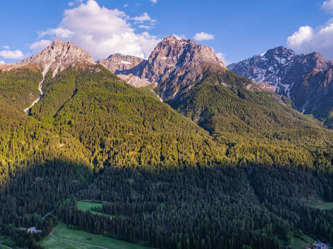 Aerial view of the majestic rocky peaks of the Engadin mountains rising above dense green pine forests under a blue sky with soft white clouds in Sent, Graubunden, Switzerland.