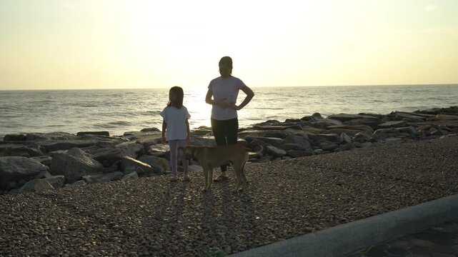 Happy mother and her young daughter standing on a rocky beach side path during sunrise while playing and waving at a friendly local dog