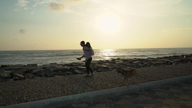 Silhouette of a loving mother giving her daughter a piggyback ride while walking on a seaside path at sunrise with a dog