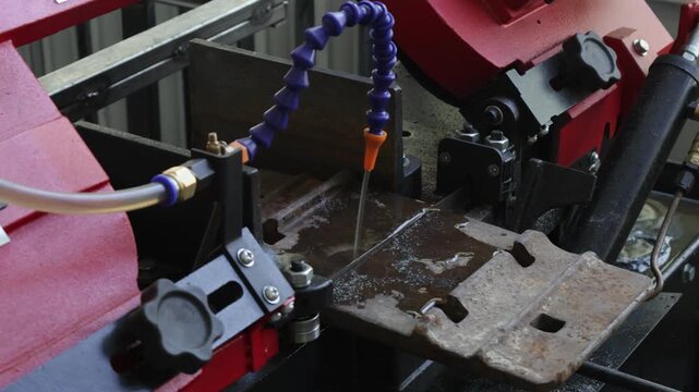 Industrial scene showing a band saw machine in action. A metal part is being precisely cut by a saw blade, while a flexible coolant nozzle delivers lubricant to the cutting zone, reducing friction and