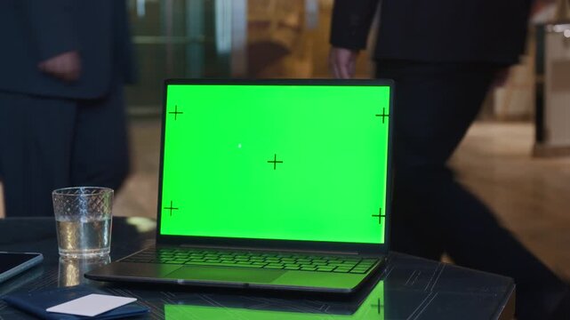 Close up of laptop with green screen and tracking markers resting on table in hotel lobby while guests passing by in blurred background. Ideal for inserting custom software interface
