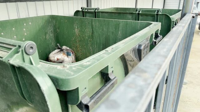 Large green dumpsters line up under a metal shelter. A single garbage bag sits inside the closest open bin. Apartment buildings stand in the background.