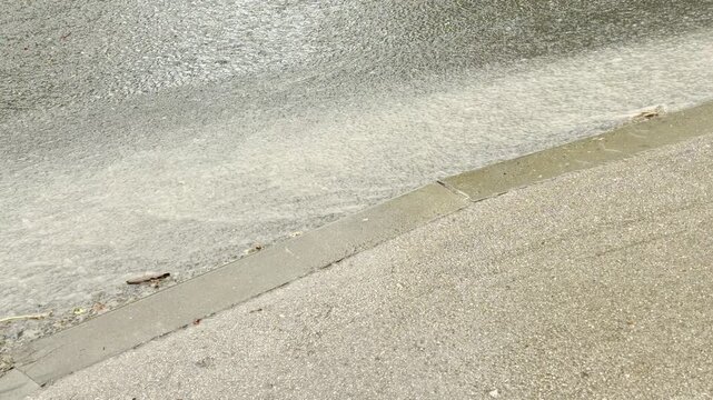 Foamy rainwater rushes down a street gutter. The murky current flows alongside a concrete curb on the wet asphalt road during a heavy downpour.