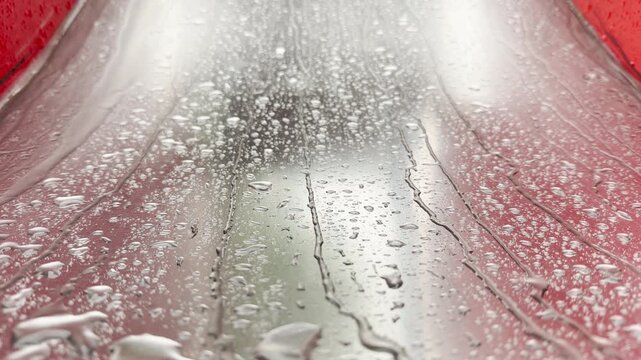 Rainwater beads up and streams down a shiny metal slide. The wet surface reflects light between bright red borders during a shower.