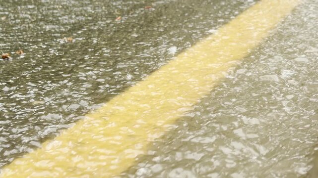 Shallow rainwater streams quickly across a yellow road marking. The water ripples over the rough asphalt surface during a heavy downpour.