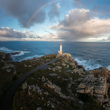 Aerial view of Faro de Punta Nariga lighthouse on a rocky headland with a vibrant rainbow arching over the Atlantic Ocean under a cloudy sky Malpica, A Coruna, Spain.
