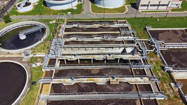 Aerial wastewater treatment plant clarifier basins showing aeration tanks, sludge channels, pipelines, circular digesters, maintenance walkways, bright midday