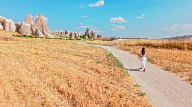 Woman tourist running along a paved path in Cappadocia, Turkey. Golden fields and fairy chimneys frame a sunny countryside travel scene.