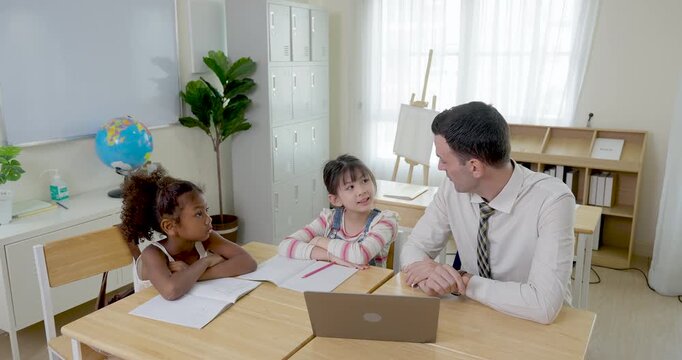Adult male teacher guides two young girls at wooden desk with laptop and notebooks, encouraging question time, peer learning, confidence, and primary classroom participation today daily activity.